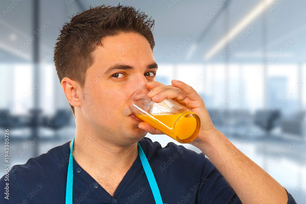 Fototapeta premium Man sips a glass of orange juice while standing in a bright modern office kitchen area, wearing a blue apron and casual shirt, conveying refreshment, health, and a short break during workday routines.