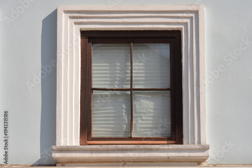 A dark wooden four pane window surrounded by a layered white stone architectural frame on a light blue building wall in bright sunlight