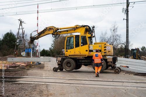 Yellow excavator on railroad tracks at a crossing, with a construction worker walking towards it