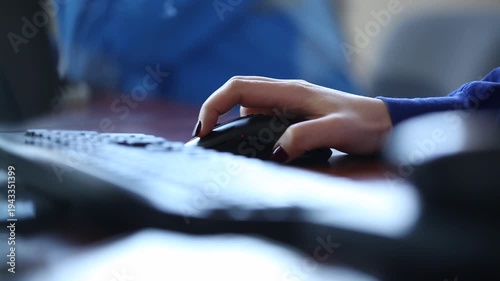 Close up shot of an unrecognizable businesswoman in a blue shirt using a black computer mouse at her office desk, with her fingers clicking next to a keyboard.