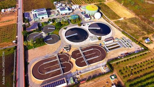 Aerial perspective of industrial water treatment infrastructure with clarifiers, solar panels, and sludge processing units on a sunny day. Circular sedimentation tanks and oxidation ditches.