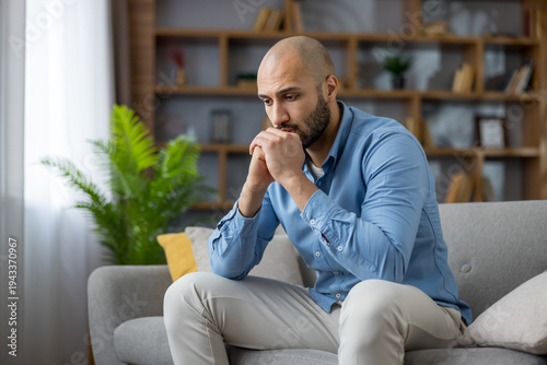 фотографія Upset young man sitting on sofa at home, looking pensive and worried, experienci