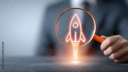 A hand holding a magnifying glass with a rocket icon, representing startup discovery, innovation scouting, and the search for high growth business opportunities.
