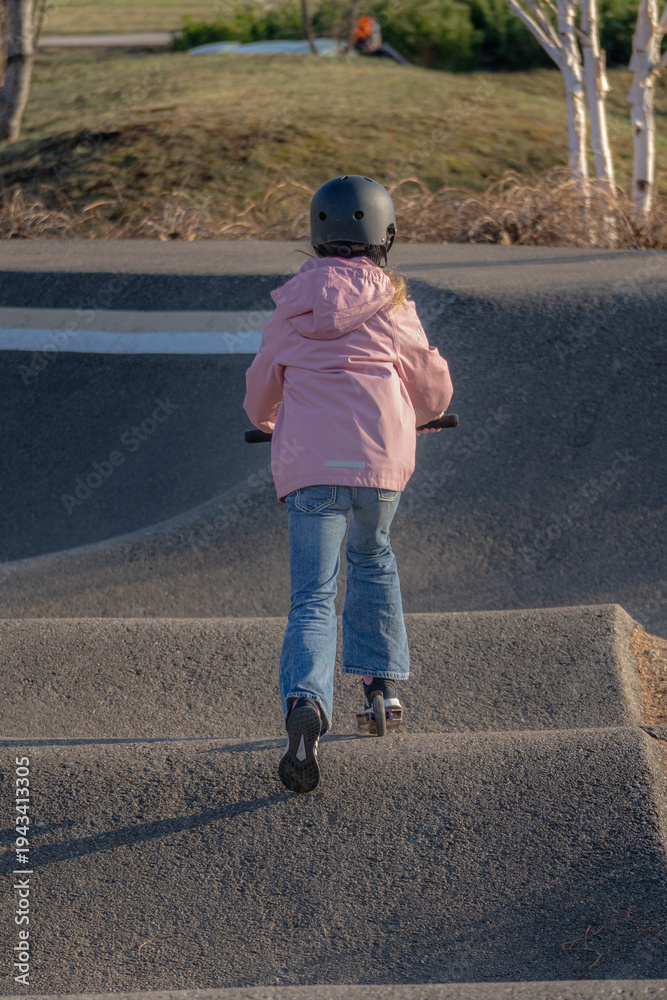 Fototapeta premium Young girl in a pink jacket and black helmet riding a scooter on a pump track at a skate park, back view, active outdoor lifestyle.