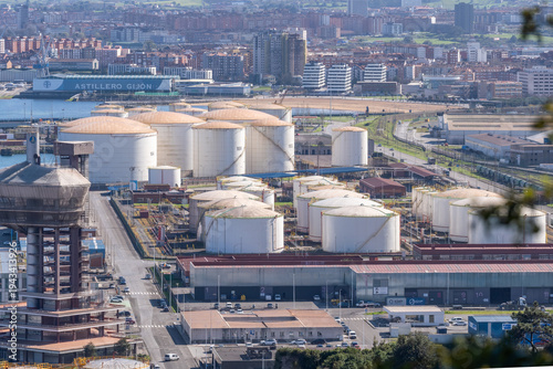 A row of massive white cylindrical storage tanks for hydrocarbons and liquid fuel at the industrial port of El Musel, Gijon, Asturias, Spain.