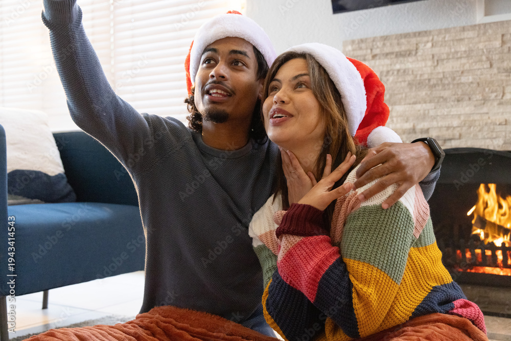 Fototapeta premium Couple sitting on blanket in front of fireplace wearing Santa hats and taking selfie