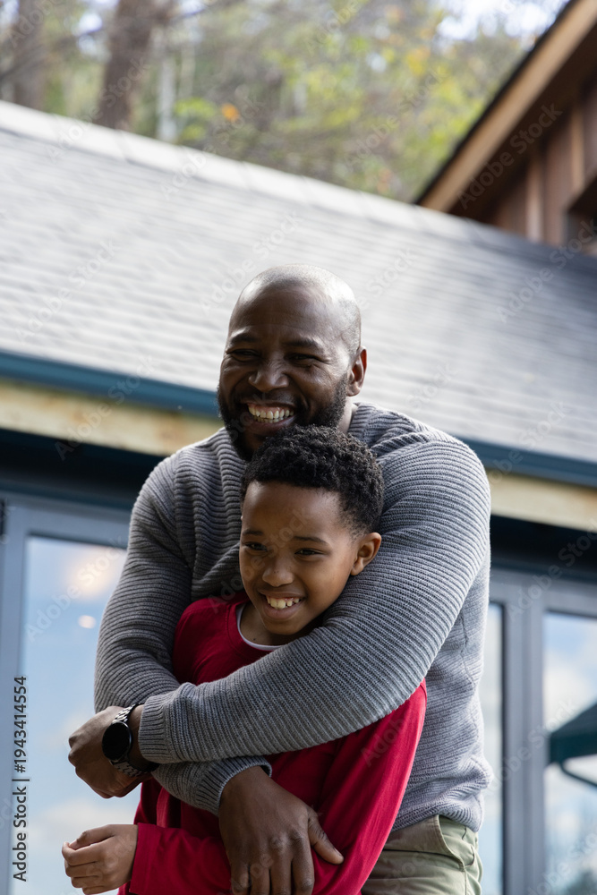 Fototapeta premium African American father hugging preteen son, smiling on porch with glass doors, showing wristwatch