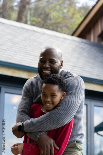 African American father hugging preteen son, smiling on porch with glass doors, showing wristwatch