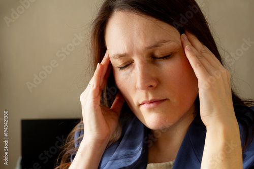 Photos Woman with brown hair touching her temples indoors, headache or stress, closed eyes, blue shirt, home environment, daylight