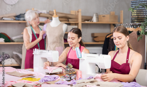 Canvastavla Two female seamstresses work on a sewing machine next to an elderly dressmaker who checks a sample of a dress on a mannequin