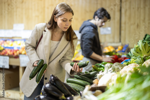 Bild auf Leinwand Positive woman chooses cucumbers for purchase in greengrocer shop