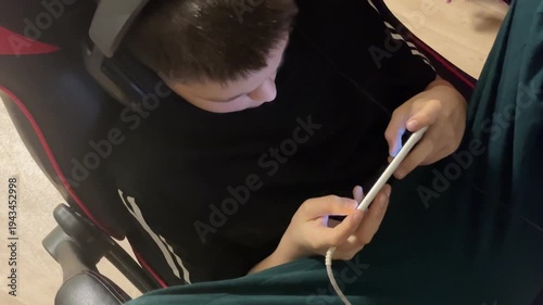A 12-year-old boy sits in the evening wearing headphones, absorbed in his smartphone, the screen lighting his focused face as he listens and plays. Mobile games.