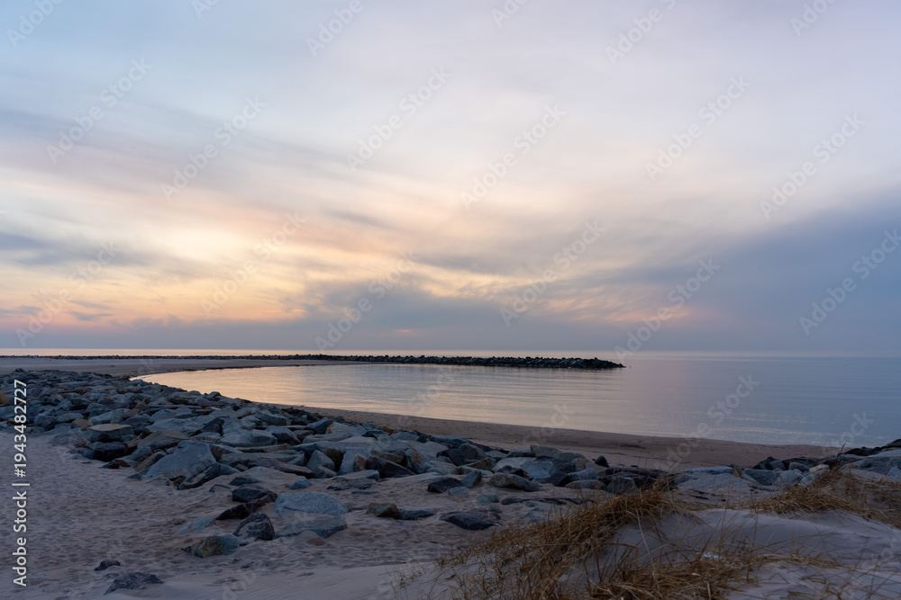 Obraz premium Deserted sandy beach with rocky breakwater curving into calm sea under soft pastel twilight sky at early winter dawn. Concept of coastal solitude, off-season travel, raw nature and peaceful emptiness.