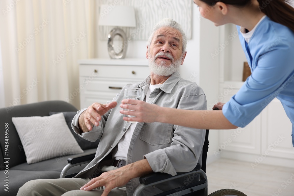 Obraz premium Care worker giving glass of water to senior man in wheelchair indoors