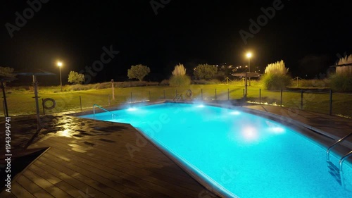 A beautiful outdoor heated swimming pool glows vibrant blue at night, with steam rising from the warm water. The tranquil scene features a wooden deck, lush green grass, and distant ambient lights