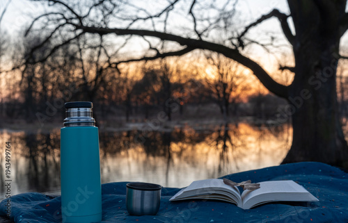 Open book and blue thermos with steel cup on blanket by calm river at golden sunset in autumn park. Concept of outdoor reading, slow living, mindful relaxation and cozy nature escape.