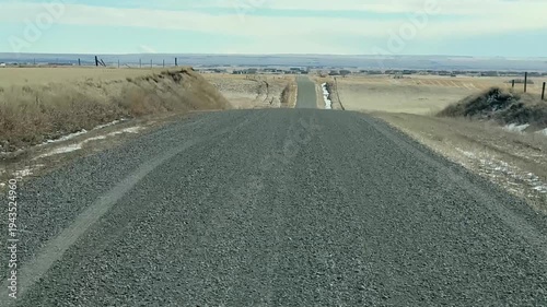 Drivers POV driving along empty gravel country road across wide prairie landscape under blue sky in Alberta Canada.
