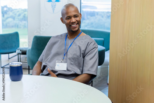 Man in gray scrubs sitting, smiling at staff lounge round table with tumbler and ID badge