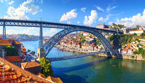A sweeping bridge arcs over a river, with a city of red-tiled roofs and colorful buildings on either side under a blue sky