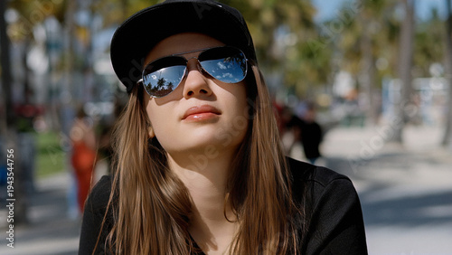 Youthful optimism and summer vibes A young woman looks upward with confidence wearing sunglasses and a baseball cap in a sunny urban outdoor setting A sense of aspiration and a bright future travel li