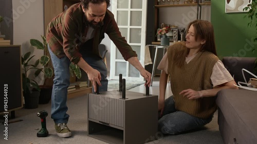 Full shot of joyous middle-aged Caucasian couple attaching legs to flat pack drawer, finishing with task, turning over, admiring work, then giving each other high fives, hugging and kissing