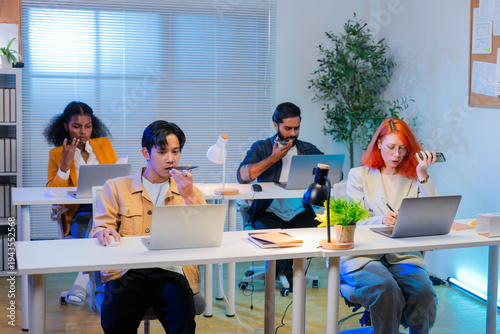 Diverse group of young professionals using smartphone voice messages while working on laptops in a modern office. Multicultural team communicating with mobile technology, collaboration tools