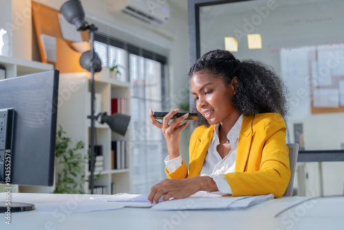 Confident professional businesswoman using smartphone voice message while reviewing documents at modern office desk. Young corporate employee working with technology, communication tools, and producti