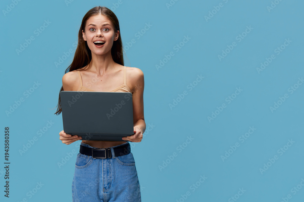Obraz premium young woman holding laptop with surprised expression, casual clothing and blue background, technology and digital concept in modern lifestyle