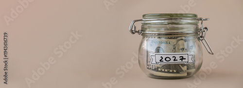 Saving Money In Glass Jar filled with Dollars banknotes. 2027 year transcription in front of jar. Managing personal finances extra income for future insecurity. Beige background