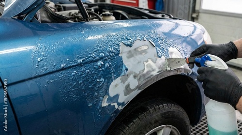 Close view of a blue car fender being repaired with sanding and filler while a technician sprays cleaner in an auto body shop, practical and focused mood