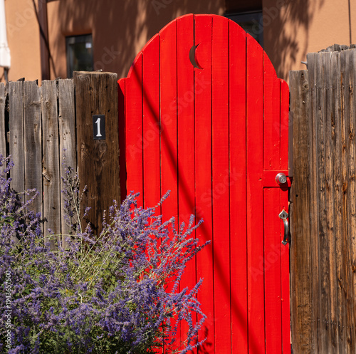 bright red wooden door with half moon cut out and diagonal shadow going through the middle in yard in home or house of Santa Fe New Mexico purple flowers in front garden in bright sun in summer time 