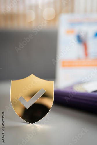 Gold shield icon with check mark on desk for insurance protection and data security concept. Guaranteed safety symbol for business privacy, financial trust, and verified cybersecurity service.
