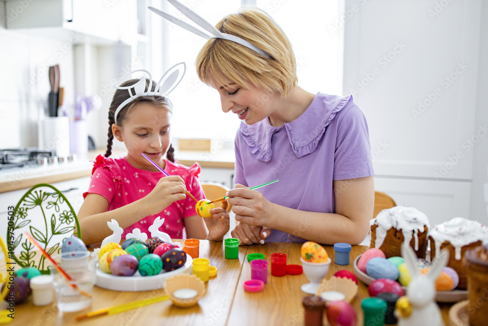 Obraz premium A mother and her daughter wearing bunny ears are happily painting colorful Easter eggs together at a wooden table, surrounded by festive decorations and cakes