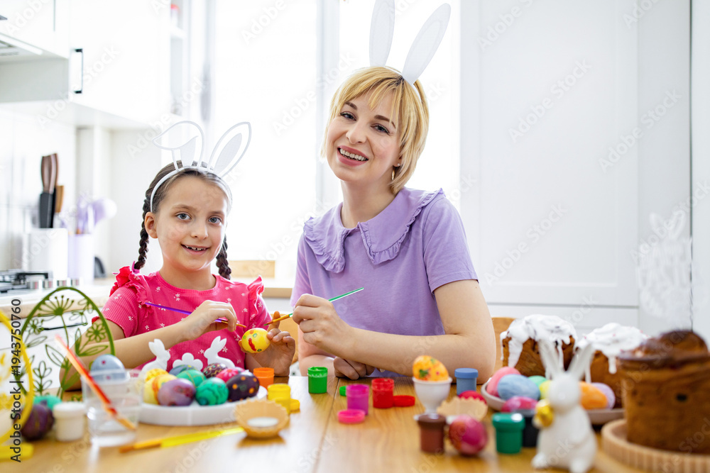 Obraz premium A mother and her daughter wearing bunny ears happily paint colorful Easter eggs together at a wooden table adorned with festive decorations and cakes