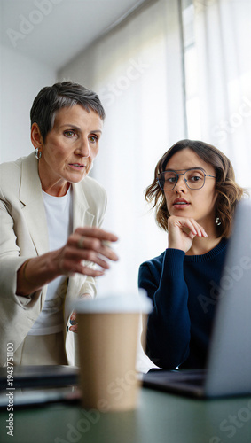 Veteran female executive intensely pinpointing a critical project flaw on the laptop screen while an attentive junior associate calmly processes the strict feedback.