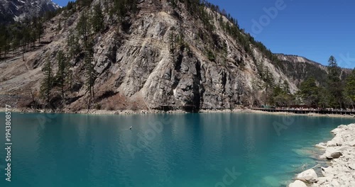 Adult man with backpack leisurely walking along wooden boardwalk beside stunning turquoise waters of mountain lake in scenic Jiuzhaigou Valley on bright, sunny day. Camera follow behind and turn left
