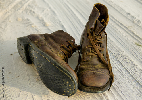 Worn brown leather boots rest on concrete ground. Frayed laces indicate heavy use. Scuffed soles prove frequent walking. Weathered leather shows age and texture. One boot faces left, the other right