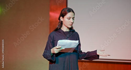 Indian young adult gen z female lady standing wear outfit dress hold script paper do play scene practice indoor hall stage. Teen age girl read story line talk speak day time college campus room place