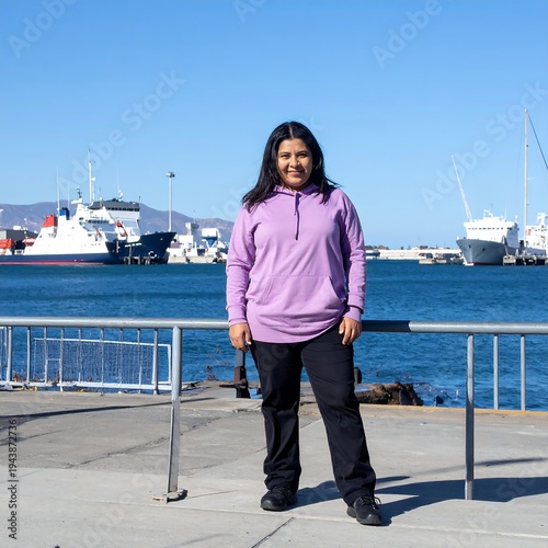 A young girl stands on a pier with a serene sea and boats behind her