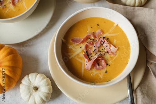 Close-up of a gourmet pumpkin cream soup with bacon, seeds, and melted cheese garnish in a ceramic bowl on a linen tablecloth with a napkin and small pumpkins.