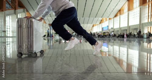Happy boy running and pushing rolling suitcase through modern Chinese railway station. Low-angle side tracking shot focusing on legs and wheeled luggage, capturing excitement and carefree joy