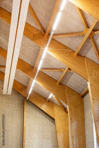 Upward view of wooden structural beams and bright ceiling lights inside a large indoor gymnasium or sports hall. Architectural timber framework and acoustic panels form a geometric ceiling pattern