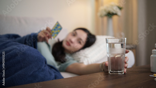 Sick young woman lying on sofa under a blanket, reaching for a glass of water while holding a blister pack of medicine. Illness, recovery, and healthcare concept.