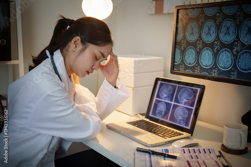 Female doctor reviewing brain MRI scans on computer screens in medical office, looking stressed and fatigued. Healthcare workload and diagnostic analysis concept.