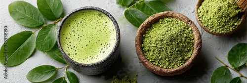 Cafe wellness aesthetic: light green matcha latte in a dark cup, surrounded by leaves and bowls of matcha powder on a textured gray surface