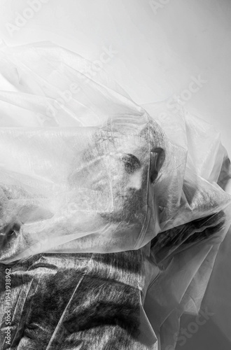 Black and white conceptual portrait of a woman partially covered with transparent plastic sheeting, suggesting themes of suffocation, confinement, or commentary against a minimal background
