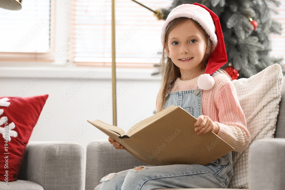 Obraz premium Cute little girl in Santa hat reading Christmas story on sofa at home