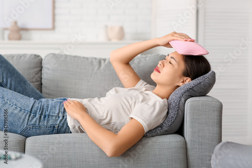 Canvas-taulu Young Asian woman warming her head with hot water bottle on sofa in living room