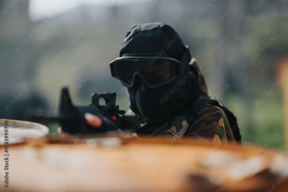 Fototapeta premium Masked paintball player in camouflage aims a marker with a scope. The scene shows a tactical shooter ready for action.