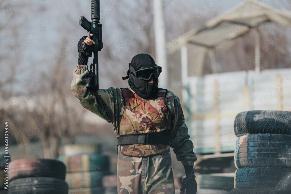 Fototapeta premium A camouflaged paintball player raises a rifle in a tactical stance, outdoors among tires and obstacles.